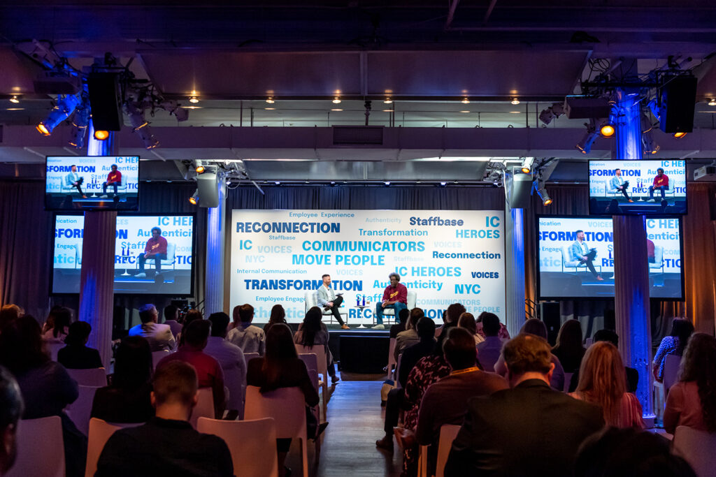 W. Kamau Bell sits on stage with host Adam Brayford at VOICES 2022 in New York City. He looks out into a packed audience and there are words in blue text highlighted behind him that say things like "Reconnection", "IC Heroes", and "Communicators Move People". 