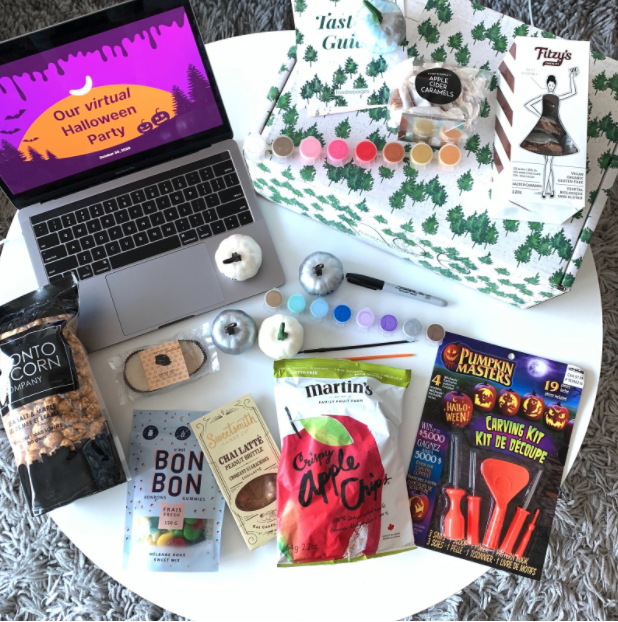 Laptop open on a table with "Our virtual Halloween party" written on the screen with spooky background. On the table there are treats, pumpkins, and a plastic carving set. 