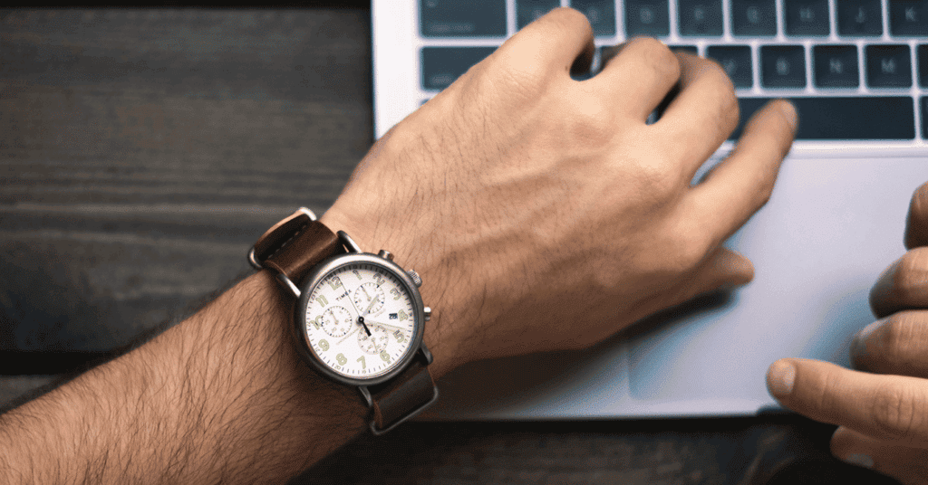 Ein Mann schaut auf seine Armbanduhr, während er am Laptop einen Social-Media-Post macht.