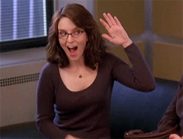 Actress Tina Fey gives herself a high five, then directs that high five to someone else by pointing at them. She wears a dark long sleeve shirt and has glasses and shoulder-length wavy hair. 