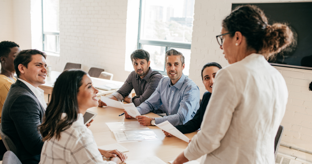 Communicating as a leader image features a woman addressing a room full of professionals in a board meeting setting