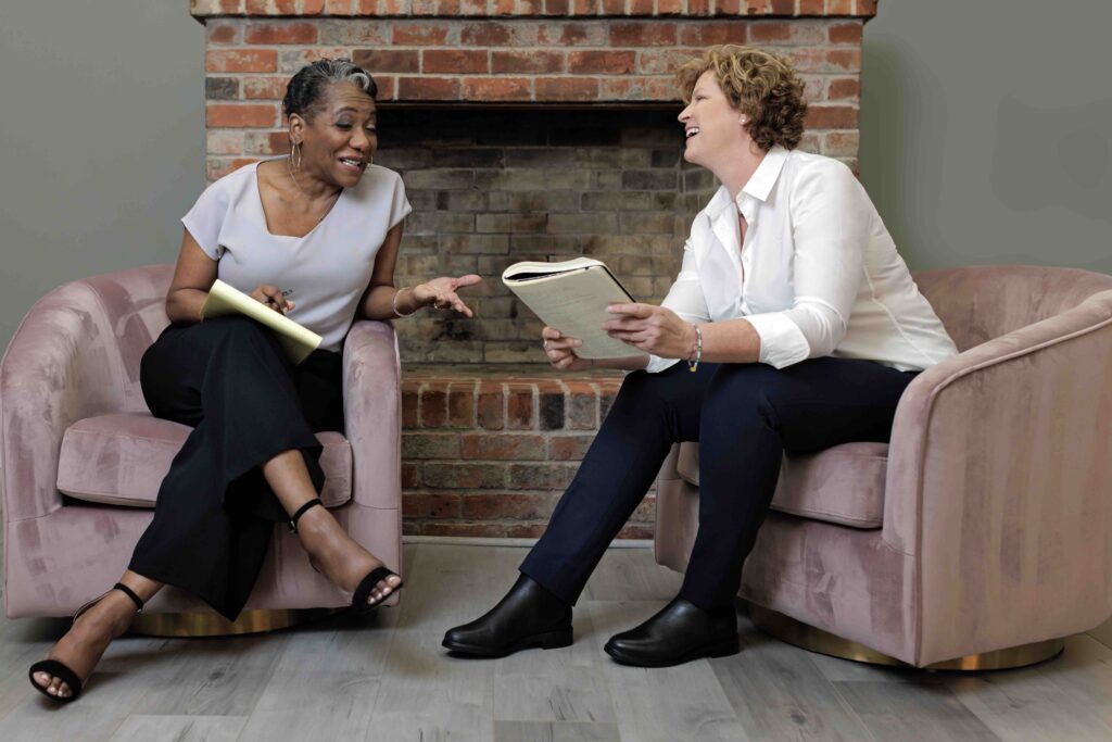 Janet M. Stovall and Kim Clark sit on two purple chairs in front of a red brick fireplace. Their books are in their hands and they share a laugh.