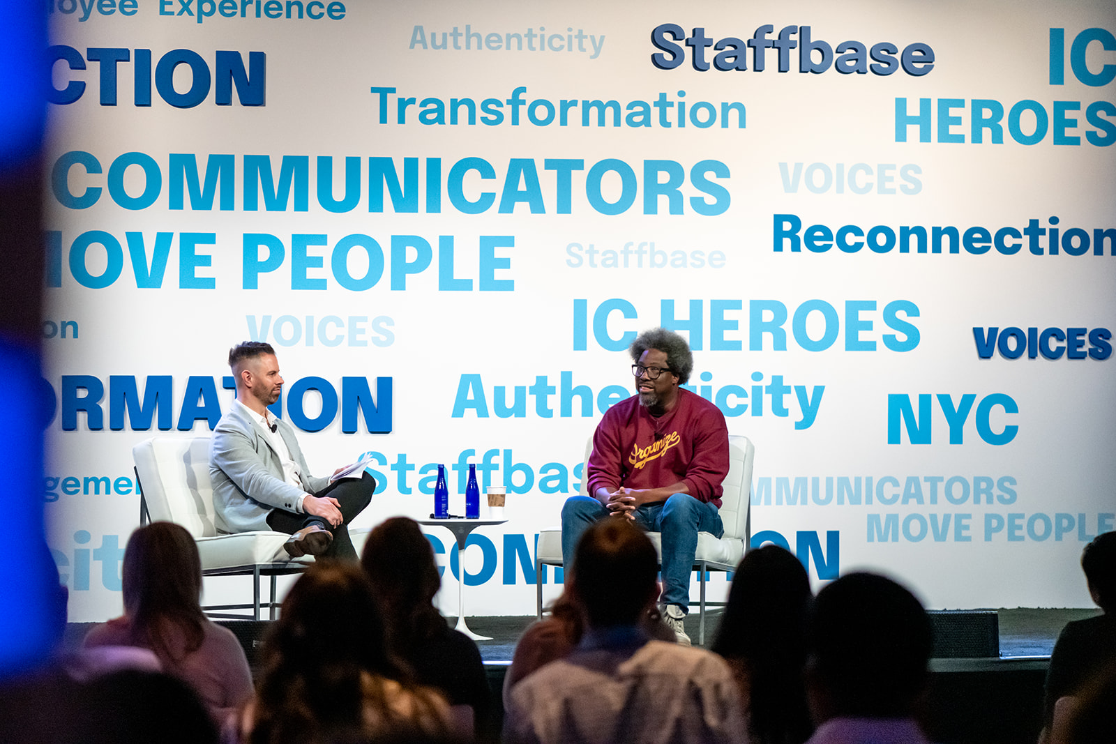 Comedian, author, and documentary producer W. Kamau Bell sits on stage with VOICES conference host Adam Brayford. They are on stage with words like "transformation" and "reconnection" and phrases like "communicators move people" in blue text behind them on a white background. We see the backs of the audiences' heads listening to Kamau and Adam.