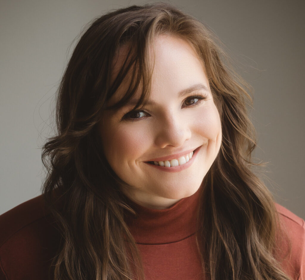 Kyla Sims, host of the Infernal Communication podcast by Staffbase, wears an orange turtlneck and smiles for the camera against a grey backdrop