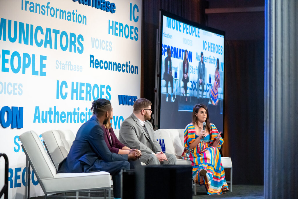 Adrienne Hoyt sits on stage with panelists at VOICES NYC. She holds a microphone and speaks to the audience.