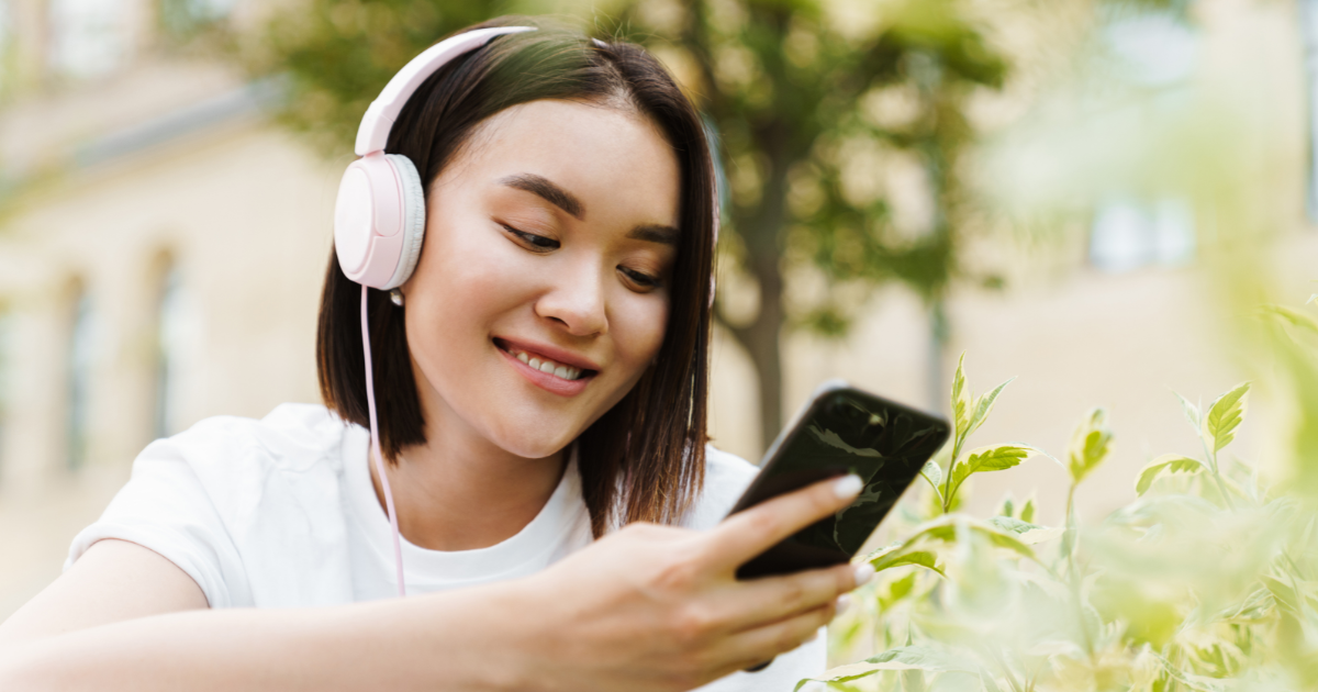 Eine lächelnde junge Frau mit kurzen Haaren trägt rosa Kopfhörer und hält ein Smartphone in der Hand, während sie draußen in einem grünen Garten sitzt. Sie scheint Musik oder einen Podcast zu hören und sieht dabei glücklich und entspannt aus.