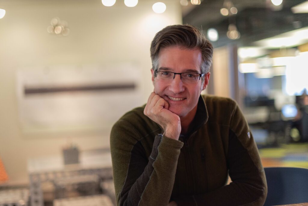 Stephen P. Anderson sits at a table and rests his head in his hand. He has dark brown and grey hair, and wears a dark green fleece sweater, black-rimmed glasses, and smiles at the camera. 