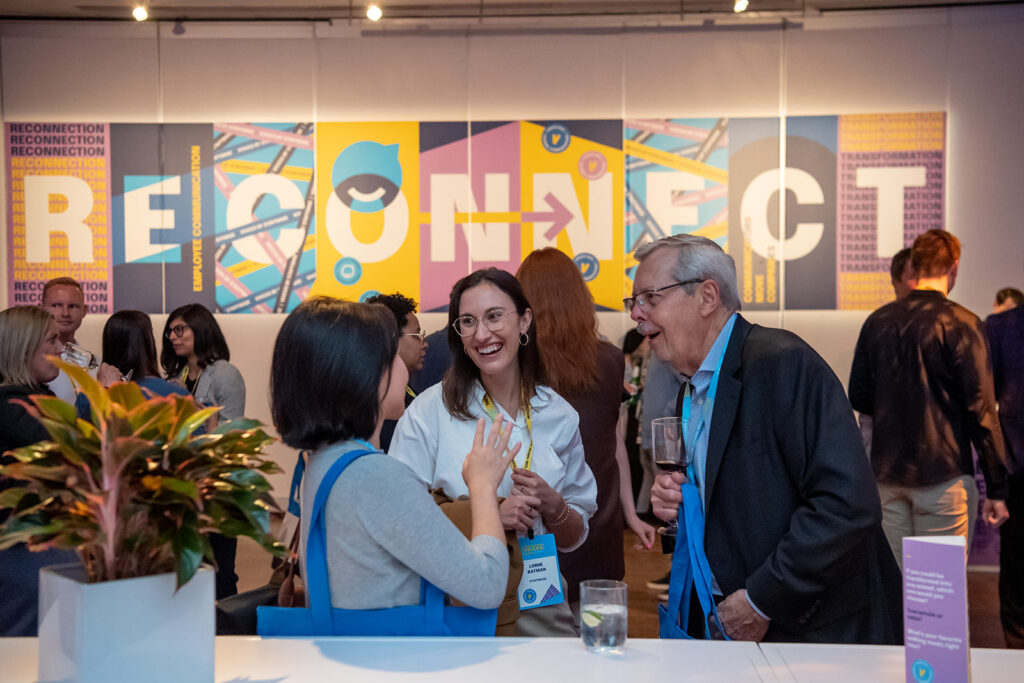 Internal communicators network at the welcome party at VOICES 2022 in New York City. A group of three people talk animatedly, smiling, and laughing with drinks in hand. Behind them are several other groups of communicators with a multi-colored sign that says "Reconnect".