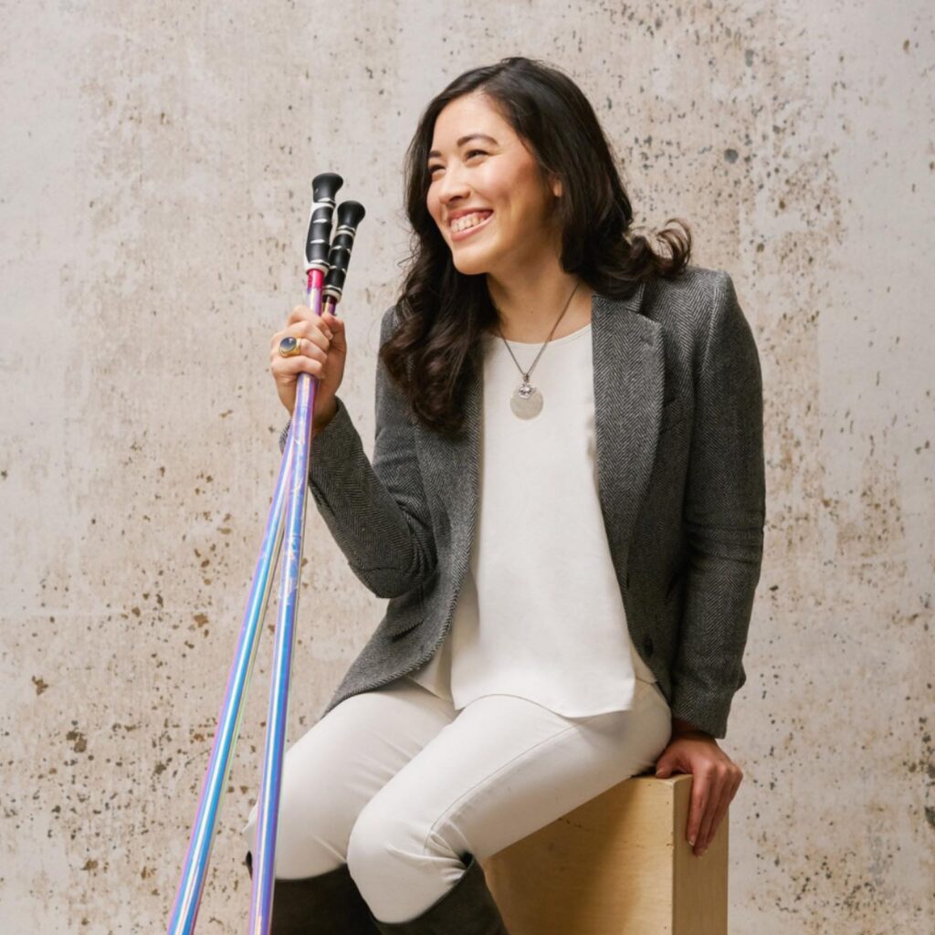 Xian Horn sits on a wooden block against a beige background. She looks to the side and smiles. She has long dark hair, and wears a grey blazer, a white tank top, a silver necklace, white pants, and black knee-high boots. She holds two walking poles that are blue and purple with black hand grips. 